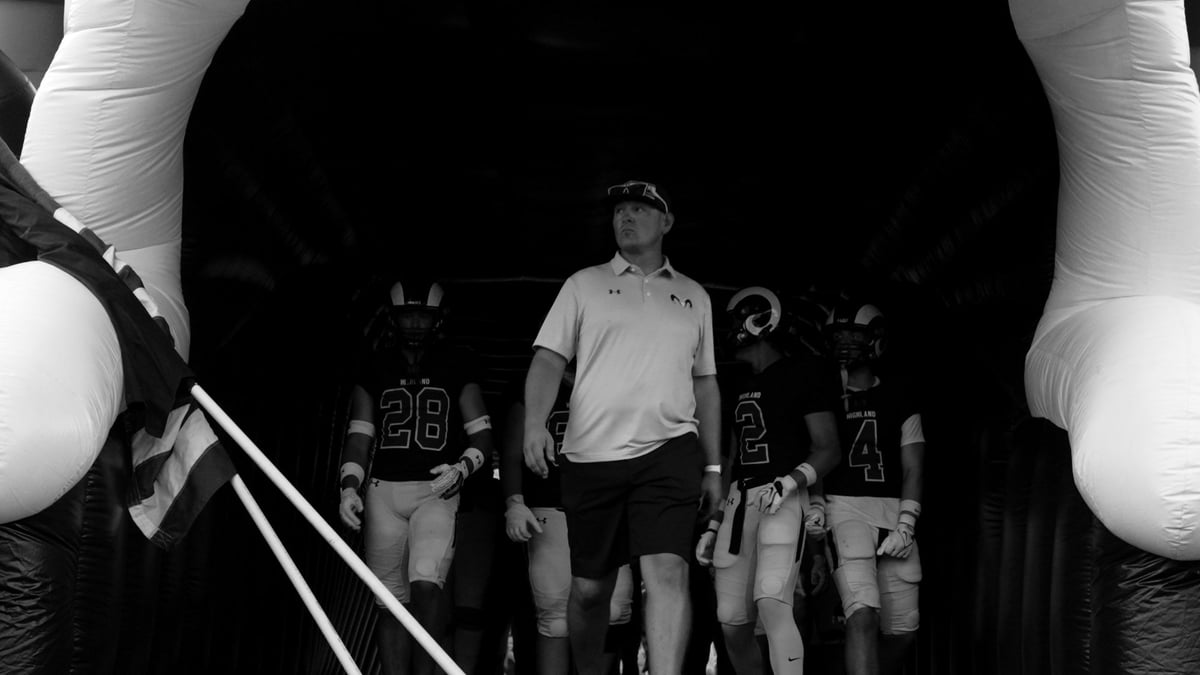 Black and white image of a coach leading a team of athletes through a tunnel with large circular lights before a game